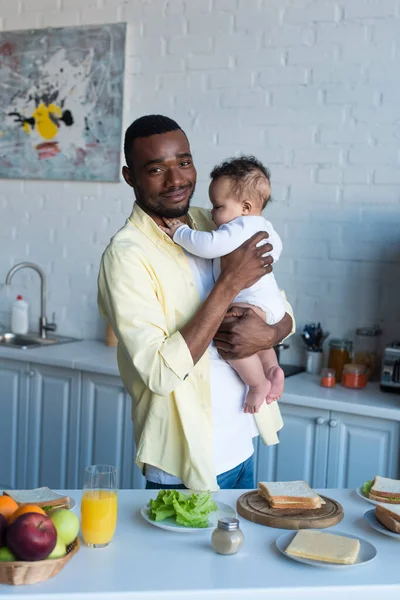 Homem americano africano sorrindo para a câmera enquanto segurando criança infantil perto da mesa com café da manhã — Fotografia de Stock