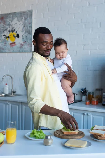 Hombre afroamericano positivo preparando sándwiches con queso y lechuga mientras sostiene a un bebé - foto de stock