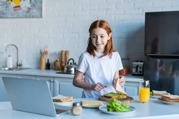 Rousse fille regardant ordinateur portable tout en faisant un sandwich pour le petit déjeuner — Photo de stock