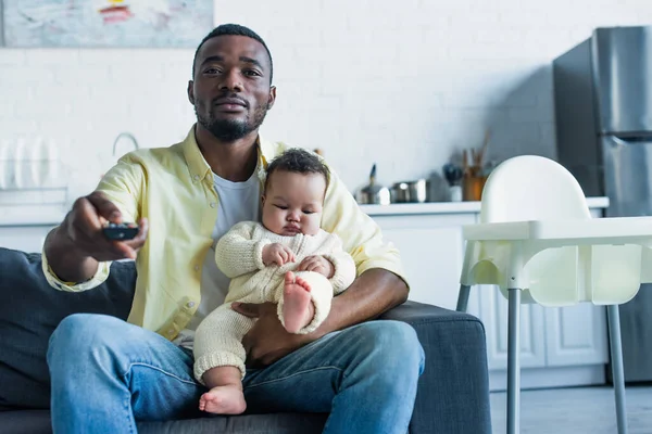 African american man clicking channels while watching tv with infant daughter — Stock Photo