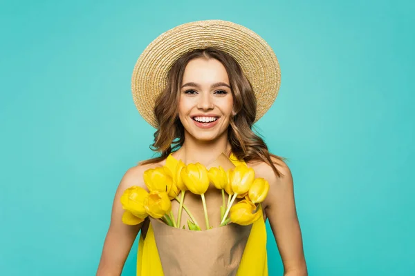 Mujer bonita en sombrero de sol sosteniendo tulipanes y sonriendo aislado en azul - foto de stock