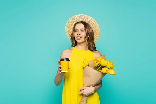 Улыбающаяся женщина в солнечной шляпе держит кофе, чтобы пойти и тюльпаны изолированы на синий — стоковое фото