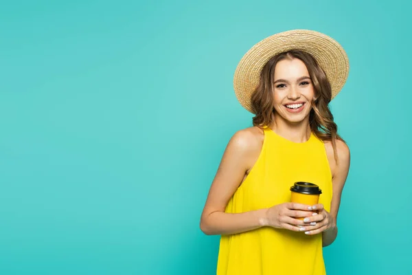 Mujer alegre en vestido amarillo y sombrero de sol sosteniendo café para ir aislado en azul - foto de stock