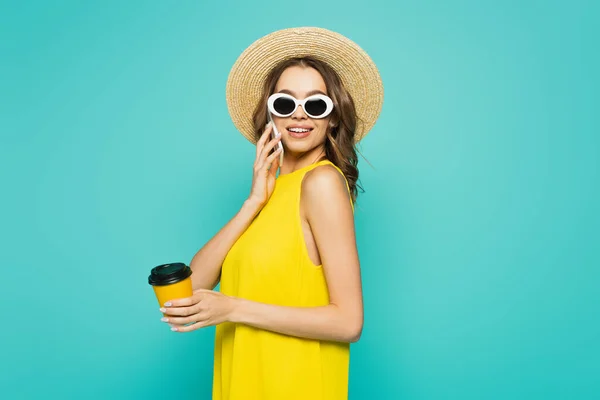 Smiling woman in sun hat talking on smartphone and holding coffee to go isolated on blue — Stock Photo