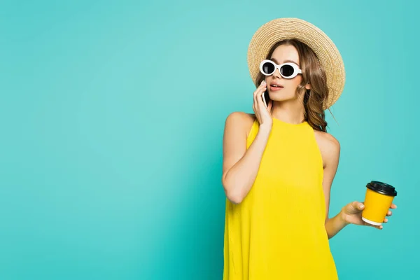 Junge Frau mit Sonnenbrille und Sonnenhut spricht mit Handy und hält Pappbecher isoliert auf blauem Grund — Stockfoto