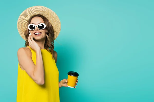 Fröhliche Frau mit Sonnenbrille hält Kaffee in Pappbecher und telefoniert auf blauem Hintergrund — Stockfoto