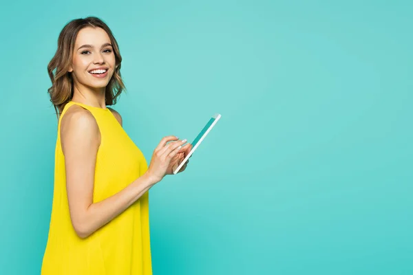 Mujer en vestido usando tableta digital y sonriendo a la cámara aislada en azul - foto de stock