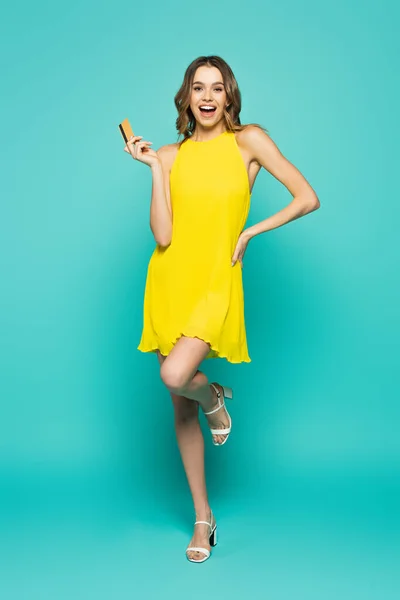 Mujer alegre en vestido con tarjeta de crédito y posando sobre fondo azul - foto de stock