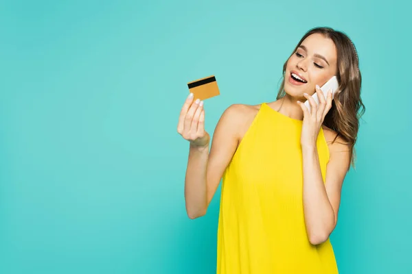 Mujer positiva con tarjeta de crédito hablando en el teléfono inteligente aislado en azul - foto de stock