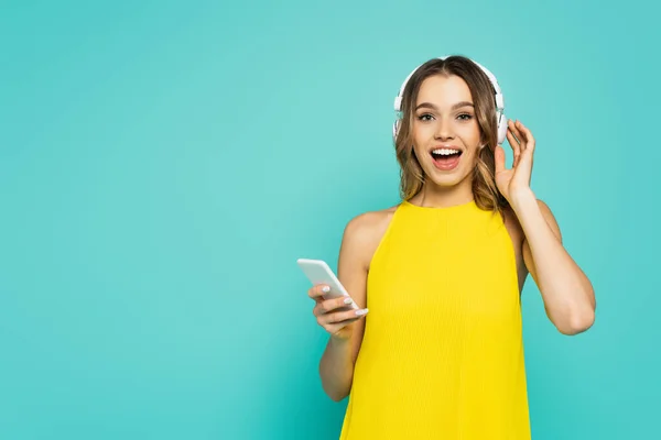 Excited woman in headphones holding smartphone isolated on blue — Stock Photo