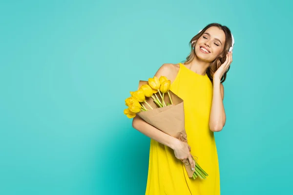 Femme souriante en robe et écouteurs tenant des tulipes sur fond bleu — Photo de stock