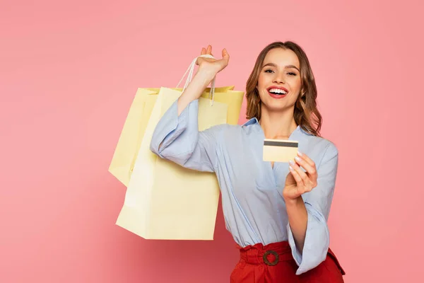 Mujer alegre sosteniendo la tarjeta de crédito y bolsas de compras sobre fondo rosa - foto de stock