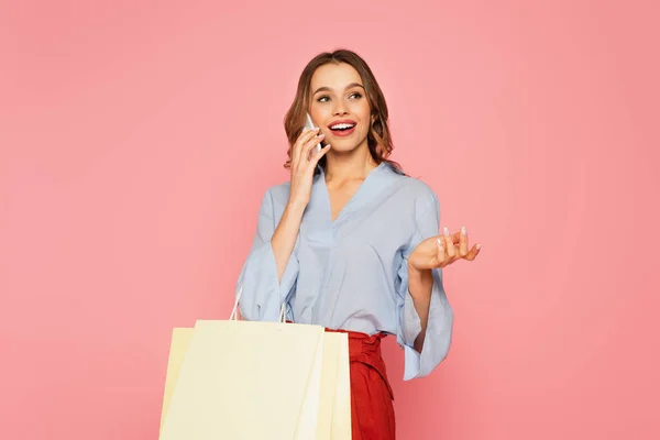 Femme souriante avec des sacs à provisions parlant sur smartphone isolé sur rose — Photo de stock