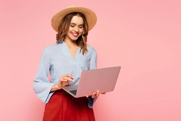 Smiling woman in straw hat using laptop isolated on pink — Stock Photo