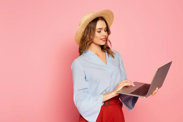 Stylish woman in sun hat using laptop on pink background — Stock Photo