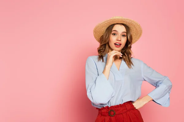 Stylish woman in sun hat holding hand on hip and looking at camera on pink background — Stock Photo