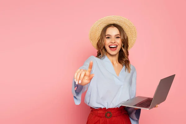 Fröhliche Frau mit Strohhut zeigt mit dem Finger und hält Laptop isoliert auf rosa — Stockfoto