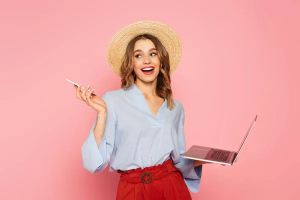 Donna sorridente in cappello solare con smartphone e laptop isolati su rosa — Foto stock