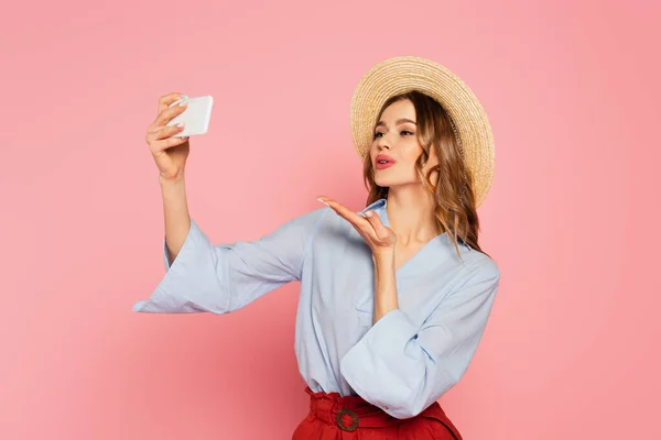 Bella donna in cappello da sole che soffia aria bacio mentre prende selfie su smartphone isolato su rosa — Foto stock