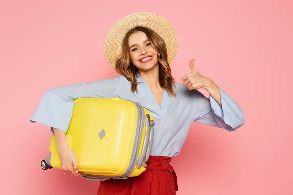 Mujer alegre en sombrero de sol sosteniendo la maleta y mostrando como gesto aislado en rosa - foto de stock