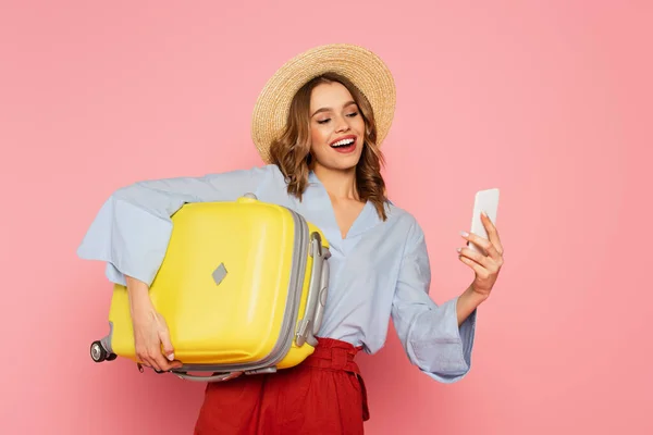 Donna sorridente con valigia utilizzando smartphone isolato su rosa — Foto stock