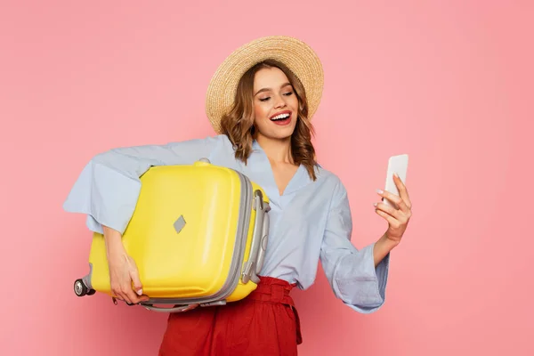 Lächelnde Frau mit Koffer mit Smartphone isoliert auf rosa — Stockfoto