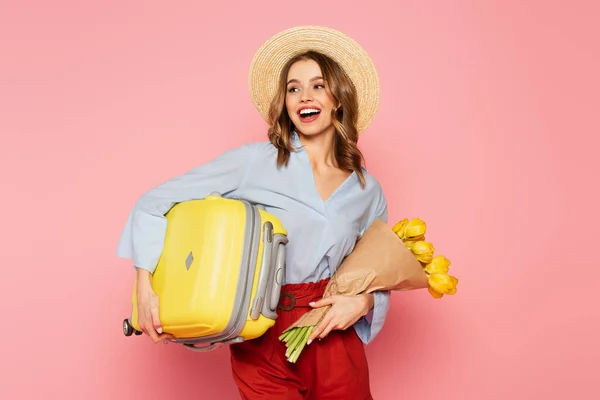 Mujer positiva en sombrero de paja sosteniendo tulipanes y maleta aislada en rosa - foto de stock