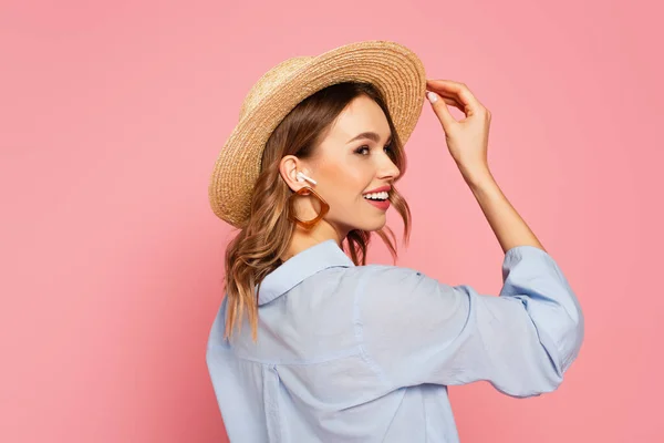 Бічний вид щасливої моделі в солом'яному капелюсі і навушниках ізольований на рожевому — стокове фото