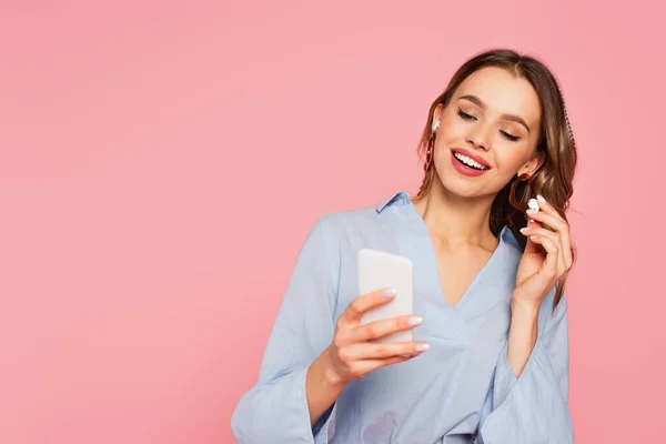 Jolie femme tenant un écouteur et utilisant un smartphone flou isolé sur rose — Photo de stock