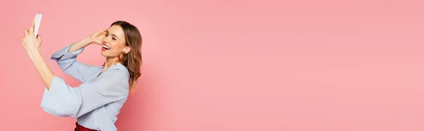 Весела жінка в навушниках має відеодзвінок на мобільний телефон на рожевому фоні, банер — стокове фото