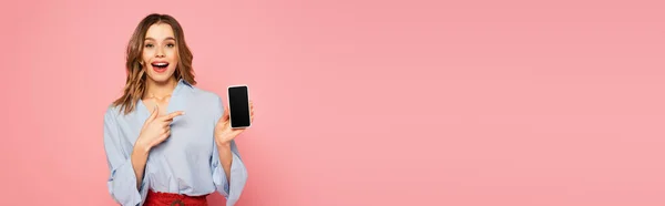 Femme excitée tenant un téléphone portable avec écran blanc et pointant du doigt isolé sur rose, bannière — Photo de stock