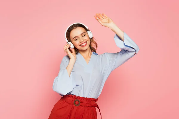 Lächelnde Frau hört Musik im Kopfhörer mit geschlossenen Augen isoliert auf rosa — Stockfoto