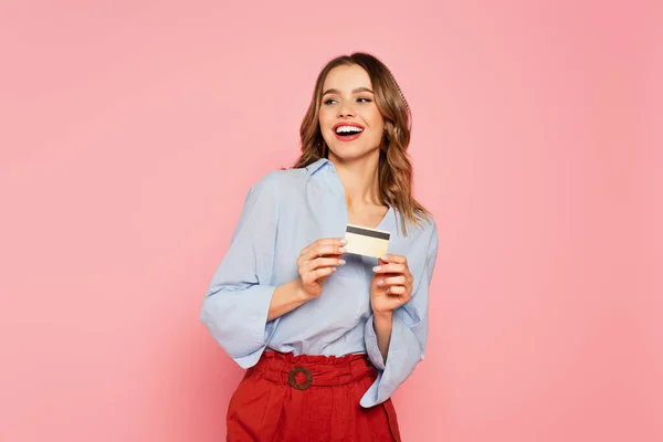 Mulher alegre com cartão de crédito olhando para longe isolado em rosa — Fotografia de Stock