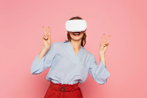 Smiling woman in vr headset showing peace signs isolated on pink — Stock Photo