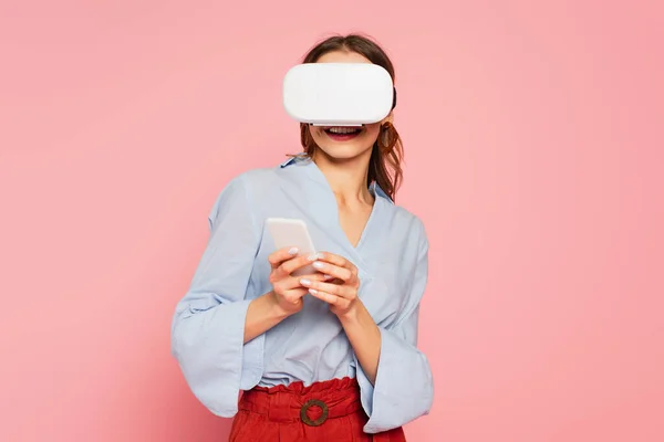 Mulher alegre em vr fone de ouvido usando telefone celular isolado em rosa — Fotografia de Stock