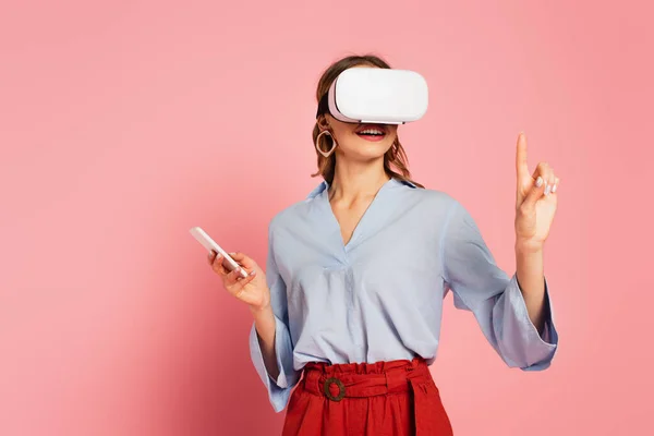 Donna sorridente in auricolare vr punta con dito su sfondo rosa — Foto stock