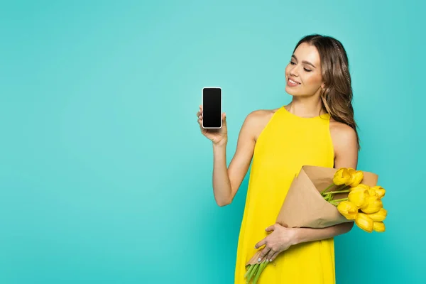 Mulher bonita em vestido amarelo segurando flores e celular com tela em branco isolado no azul — Fotografia de Stock