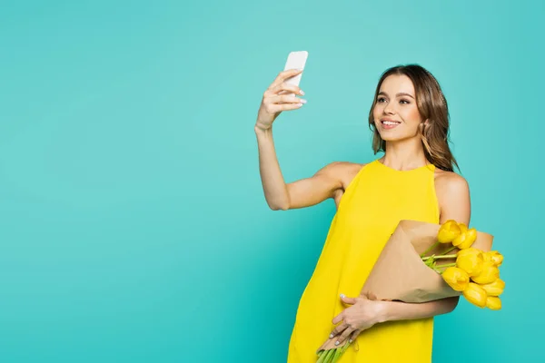 Mujer feliz con tulipanes amarillos hablando selfie en el teléfono inteligente aislado en azul - foto de stock