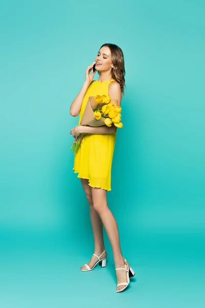 Mujer bonita con tulipanes hablando en el teléfono inteligente sobre fondo azul - foto de stock