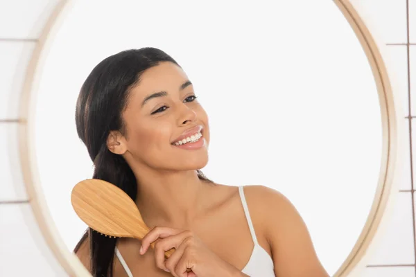 Sonriente joven afroamericana mujer cepillando el pelo cerca del espejo en el baño - foto de stock