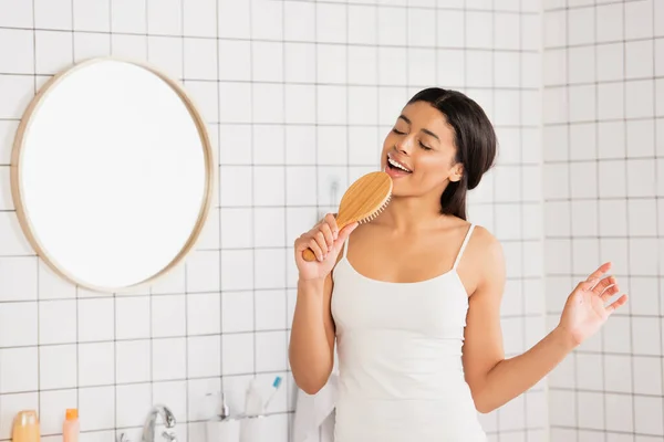 Joven mujer afroamericana en ropa blanca cantando con cepillo de pelo y haciendo gestos con la mano en el baño - foto de stock