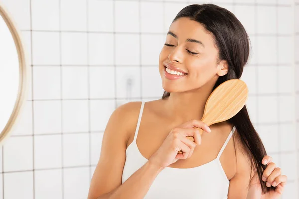 Sorridente giovane donna africana americana spazzolare i capelli con gli occhi chiusi vicino allo specchio in bagno — Foto stock