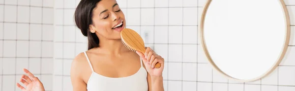 Young african american woman with closed eyes singing with hair brush in bathroom, banner — Stock Photo