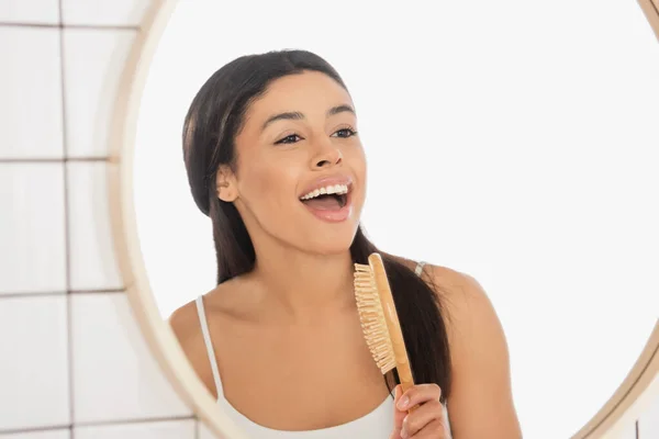 Mujer joven afroamericana cantando con cepillo de pelo cerca del espejo en el baño - foto de stock