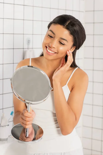 Sonriente joven afroamericana mujer en blanco tanque superior mirando en el espejo y tocando la cara con la mano en el baño - foto de stock