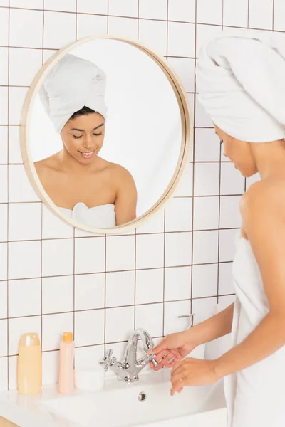 Mujer afroamericana joven envuelta en toallas de pie cerca del lavabo y tocando el grifo de metal en el baño - foto de stock