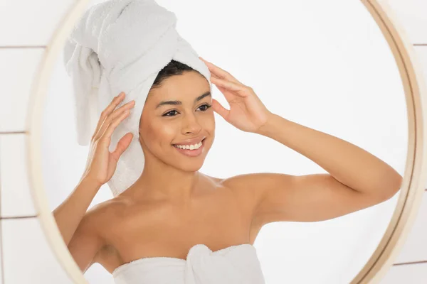 Joven mujer afroamericana envuelta en toallas cogidas de la mano cerca de la cabeza y mirando en el espejo en el baño - foto de stock