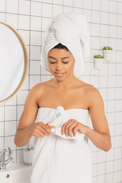 Jeune femme afro-américaine enveloppée dans des serviettes tenant brosse à dents et dentifrice près du miroir dans la salle de bain — Photo de stock