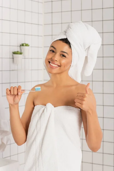 Lächelnde junge afrikanisch-amerikanische Frau in Handtücher gehüllt, Zahnbürste in der Hand und zeigt ok Geste im Badezimmer — Stockfoto