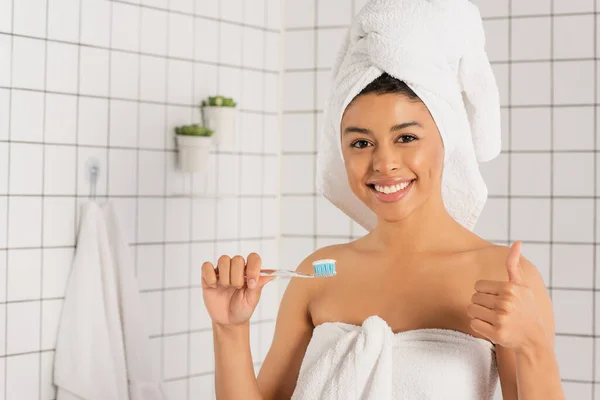 Junge afrikanisch-amerikanische Frau in Handtücher gewickelt hält Zahnbürste und zeigt ok Geste im Badezimmer — Stockfoto
